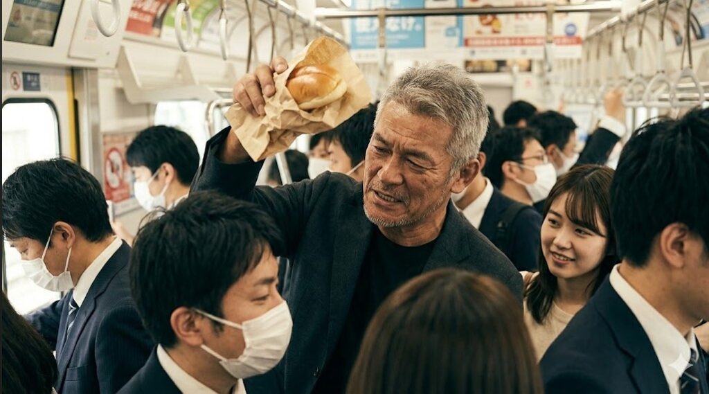 満員電車の混雑の中で、グレーのジャケットを着た短髪のベテラン日本人レスラーがコッペパンの袋を頭上に掲げて守ろうとしているユーモラスな様子